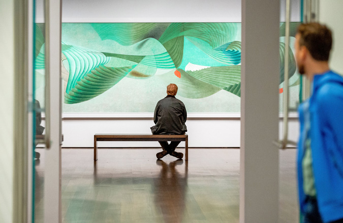 A visitor sits on a bench in front of a large, predominantly green painting in a gallery.